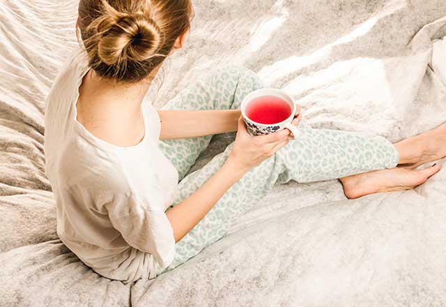 Woman holding tea cup while seated resting on bed